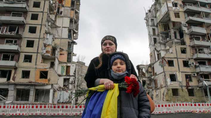 Eine Frau und ein Junge stehen mit einer ukrainischen Flagge vor einem zerstörten Gebäude.